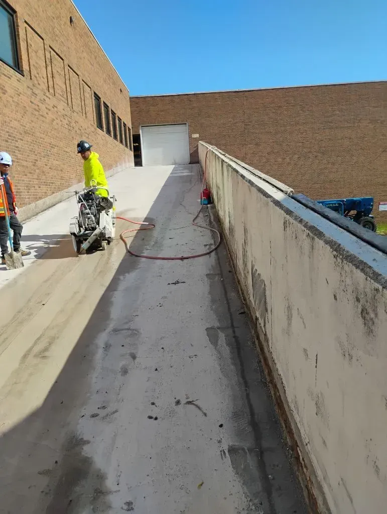 Workers using a saw to cut concrete on a ramp next to a brick building.