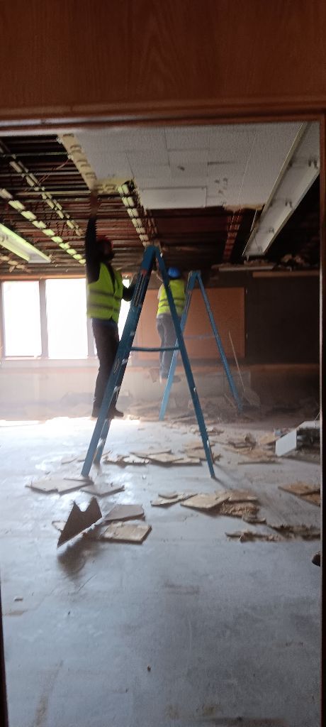 Two workers in safety vests remove ceiling tiles, standing on a ladder inside a room.