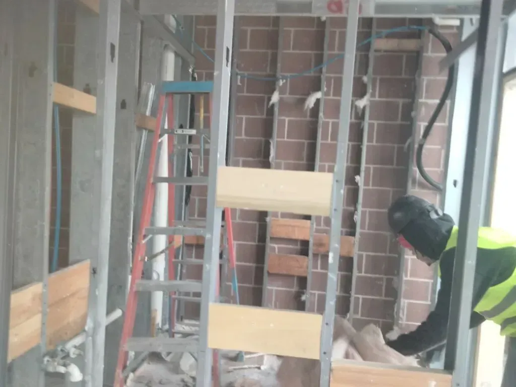 Construction worker welding metal frame in a room, with brick wall and exposed pipes visible.
