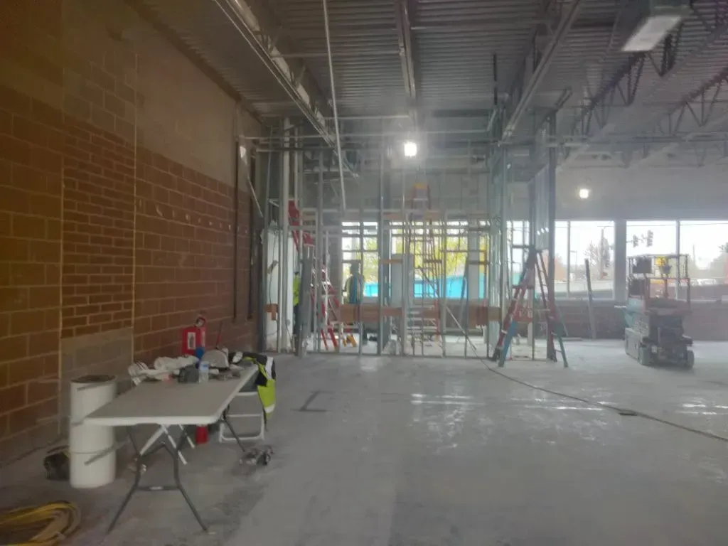 Construction site with exposed brick wall, metal framing, and concrete floor. Workers are visible.