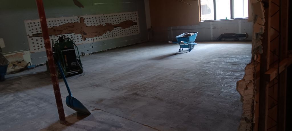 Room under construction, bare floor, damaged walls, construction tools visible, and a window in the distance.