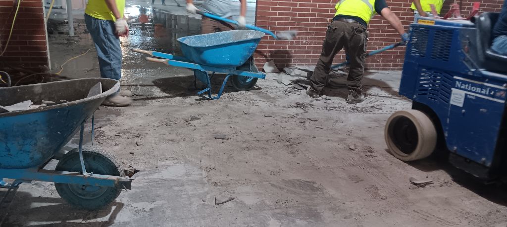 Construction workers using wheelbarrows and equipment on a concrete floor near a brick wall.