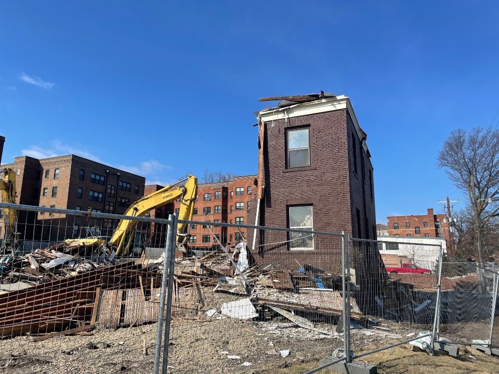 Demolition site with brick building fragment, excavator, debris, chain-link fence, and residential buildings on a sunny day.