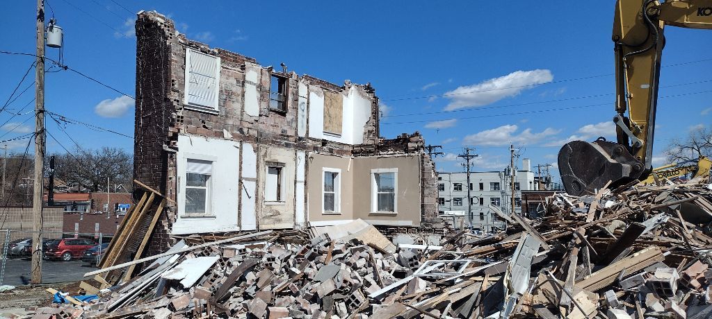 A building partially demolished, rubble in foreground. An excavator and blue sky are visible.