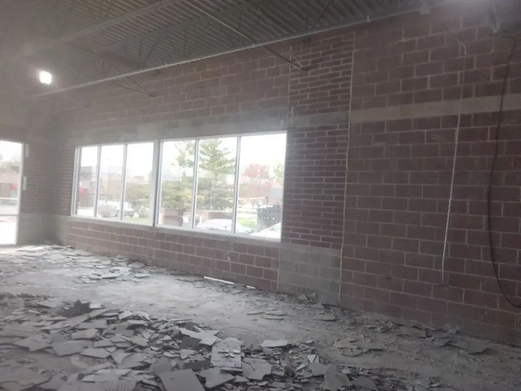 Interior view of a building under construction with exposed brick walls, large windows, and debris on the floor.
