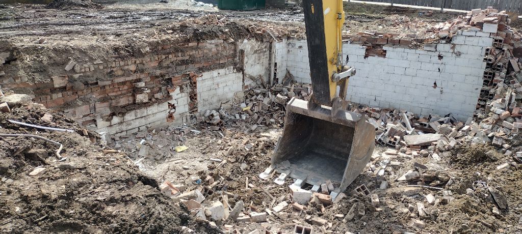 Excavator demolishing a brick structure, debris scattered, outdoors.