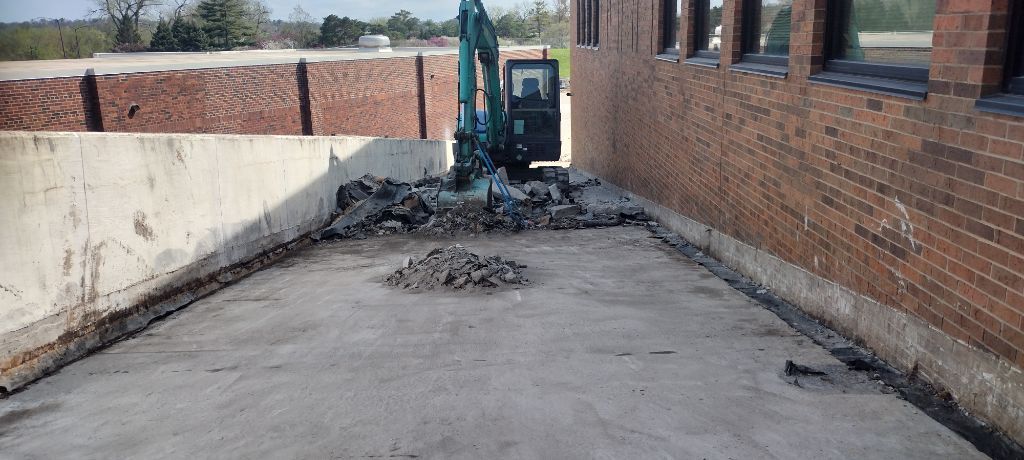 An excavator demolishes concrete next to a brick building. Gray concrete and debris fill the space.