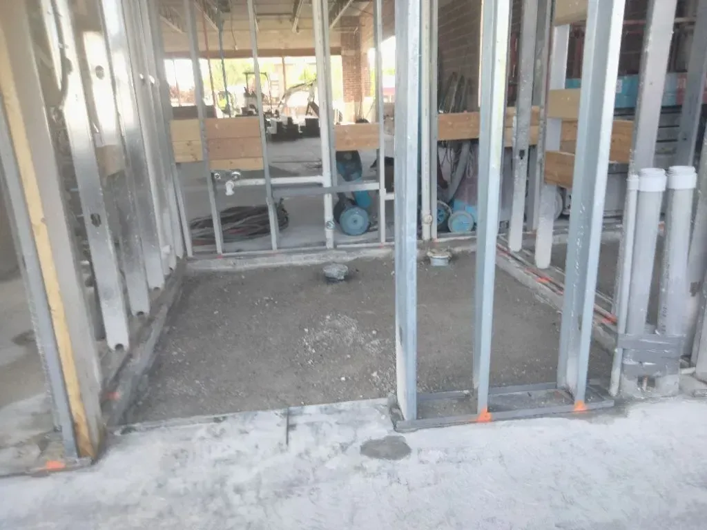 Construction site: metal framing, unfinished floor, plumbing, and equipment in the background.