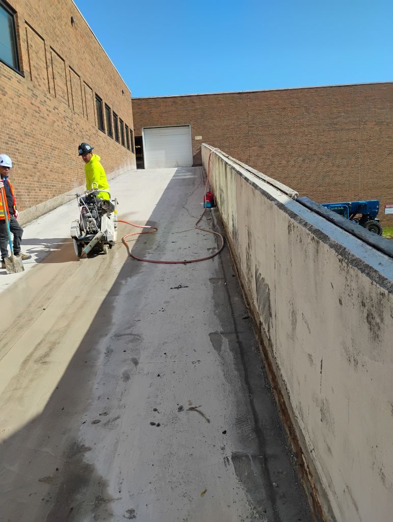 Workers cutting concrete ramp with saws near a brick building.
