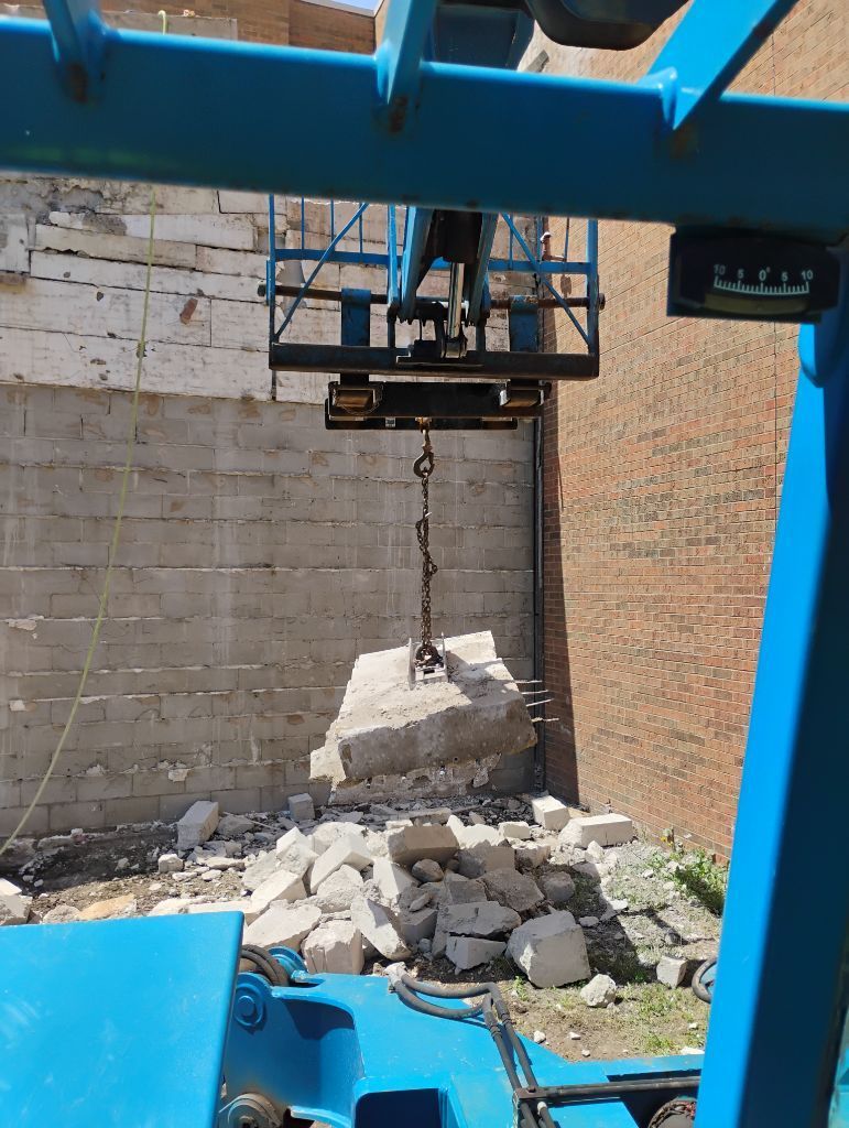 A crane lifting a large concrete block from a pile of debris near a light-colored wall.