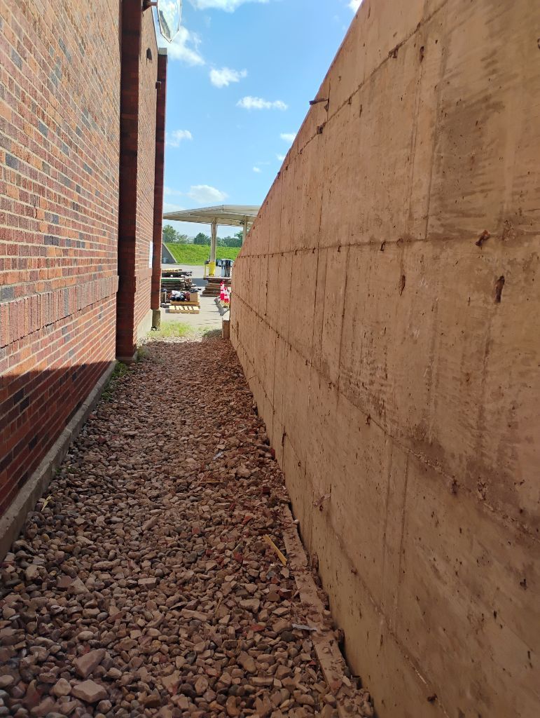 Narrow gravel path between a red brick building and a tan concrete wall, under a bright blue sky.