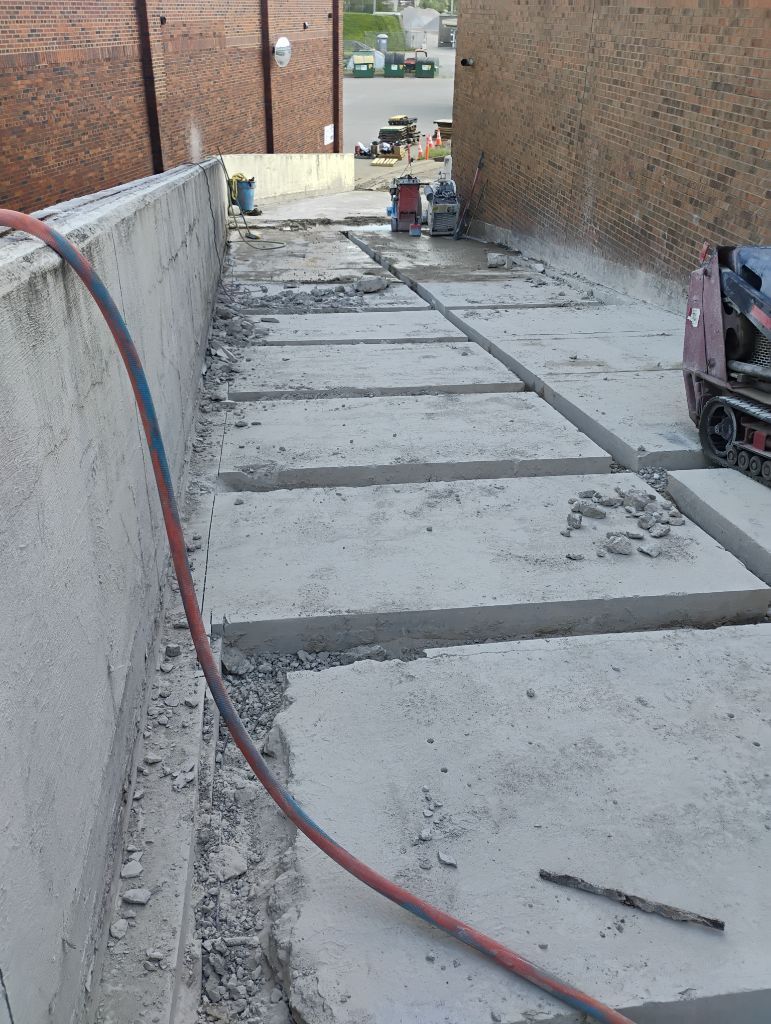 Concrete walkway being cut up for repairs. Brick buildings flank the narrow passage. Red hose on the left.