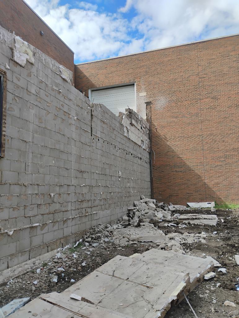 Damaged brick and cinder block exterior wall with debris, adjacent to a brown brick wall. Overcast day.