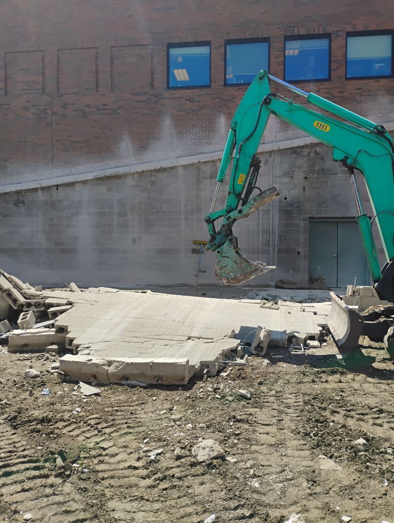 A teal excavator demolishes a concrete surface in front of a brick building.