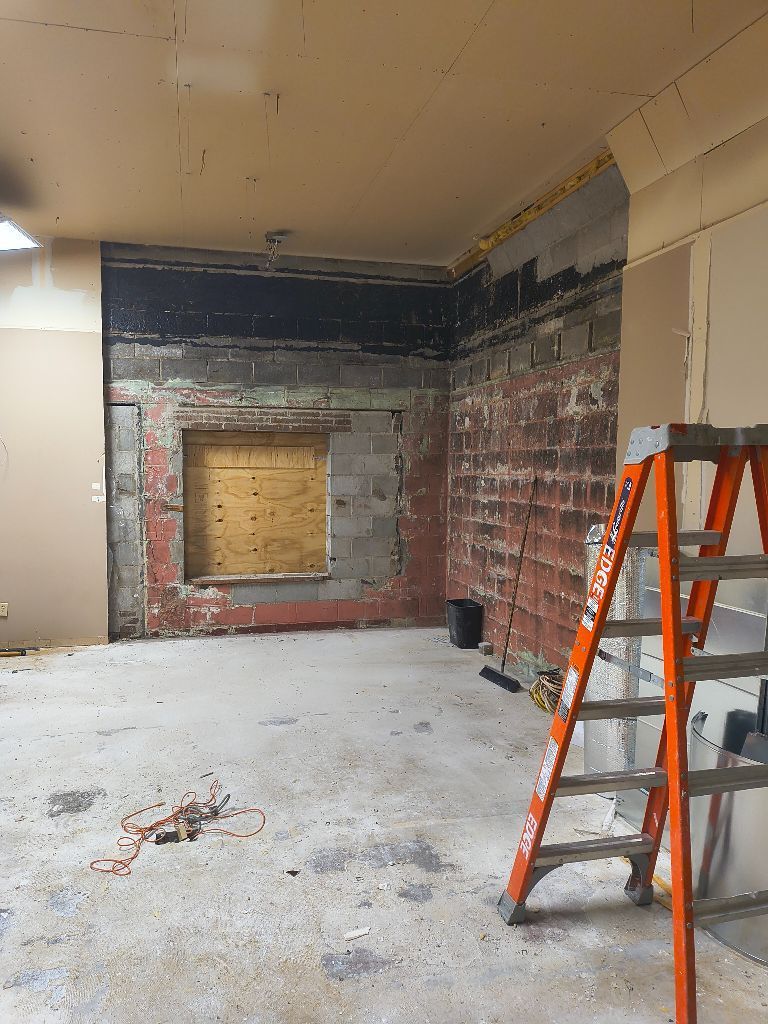 Room undergoing construction with exposed brick, plywood-covered window, and orange ladder.