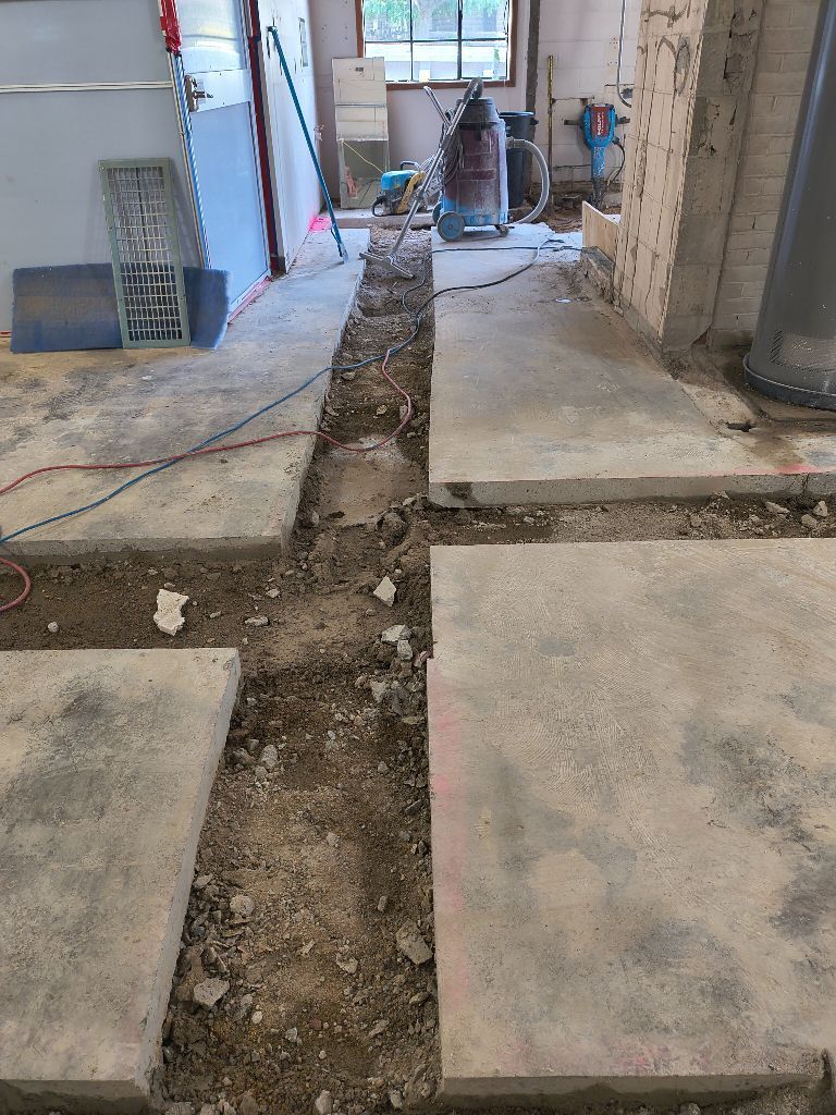 Construction site: trench dug in concrete floor, tools and materials visible.