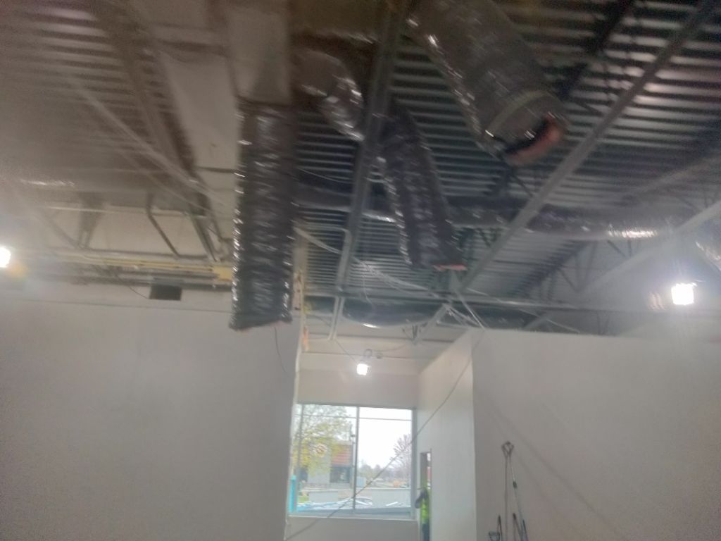 Interior view of a building under construction. Ductwork and ceiling supports are visible. White walls and a window in the distance.