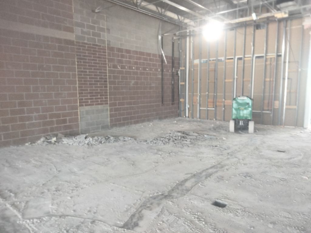 Construction site interior with brick and metal walls, concrete floor, and a small green machine.