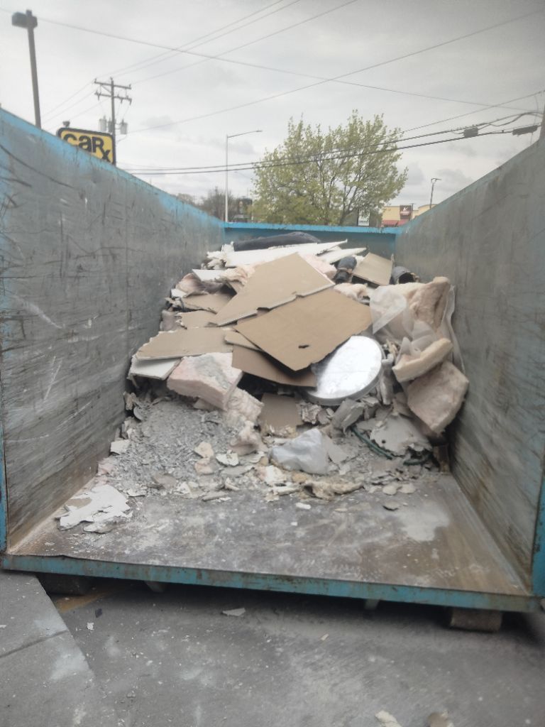 Construction debris in a blue dumpster, containing drywall, cardboard, and other materials.
