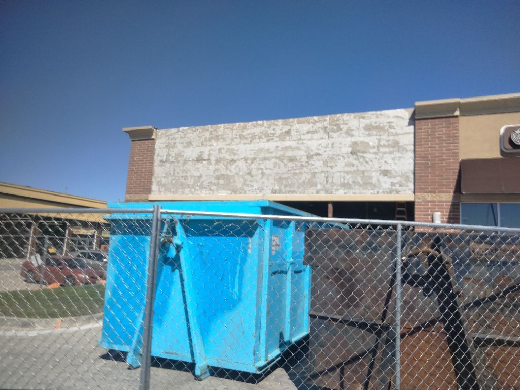 Blue dumpster in front of a building being renovated, behind a chain link fence on a sunny day.