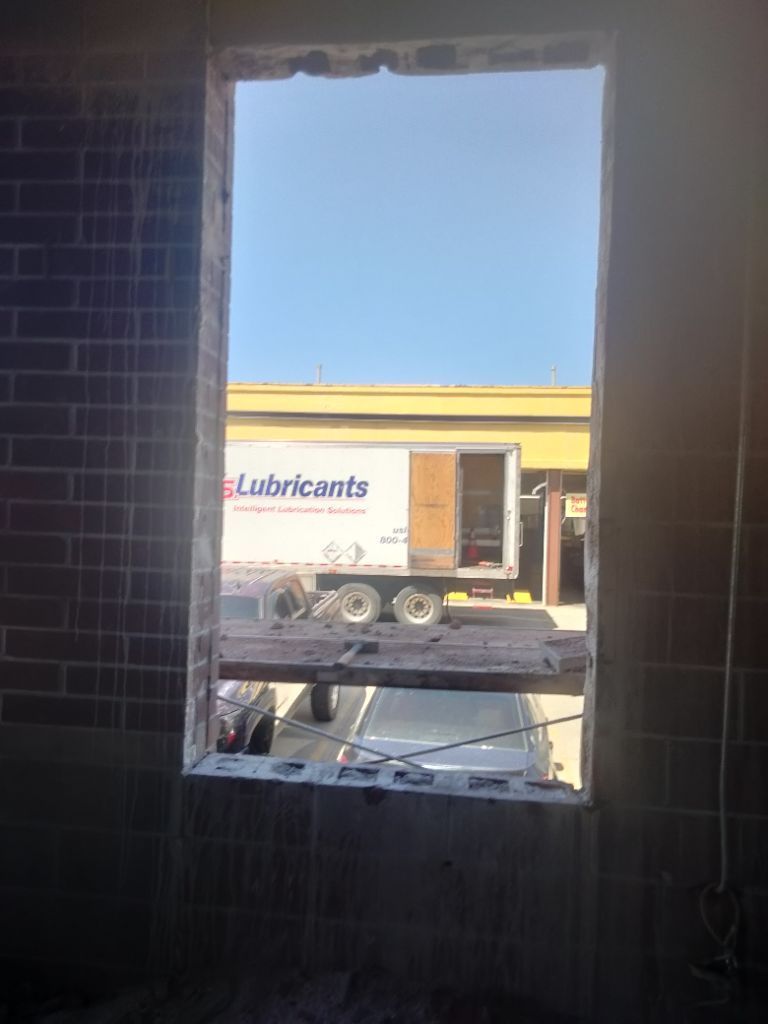 View through an unfinished rectangular opening in a brick wall, looking out at a semi-truck with a white trailer under a blue sky.