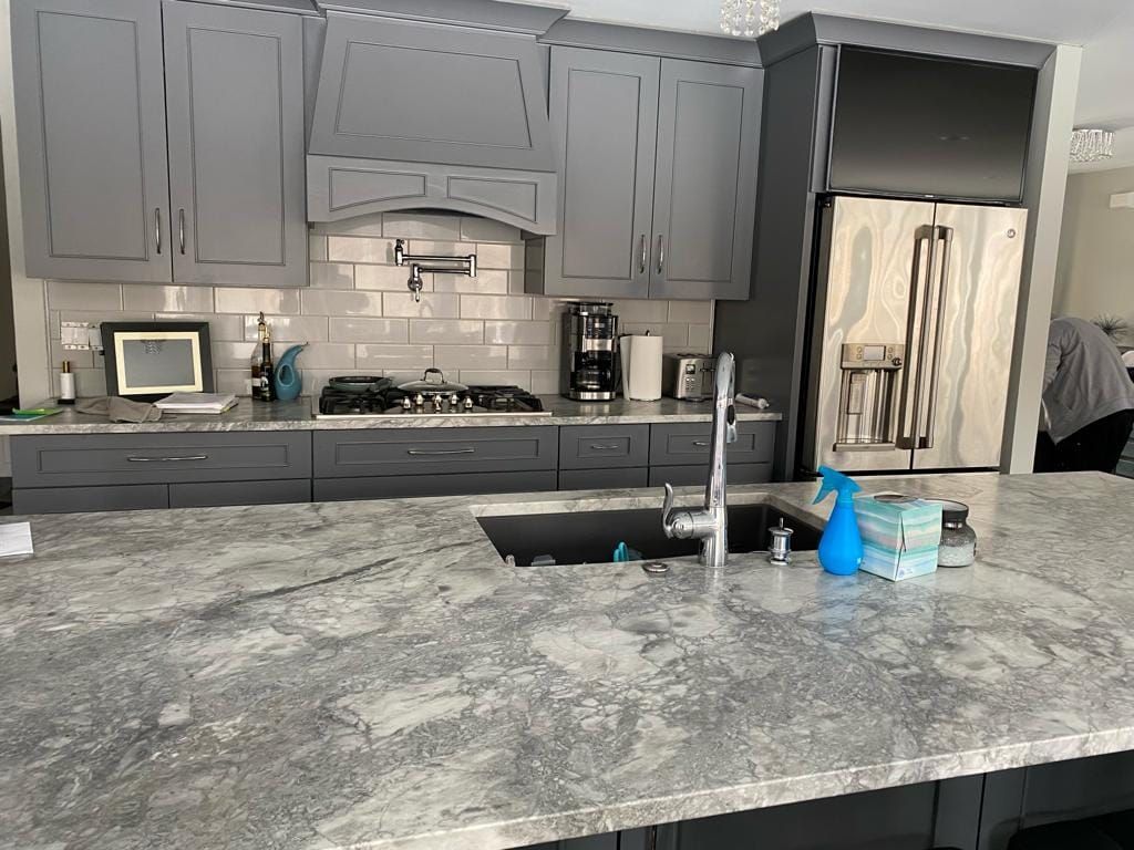 A kitchen with gray cabinets , granite counter tops , a sink , and a refrigerator.