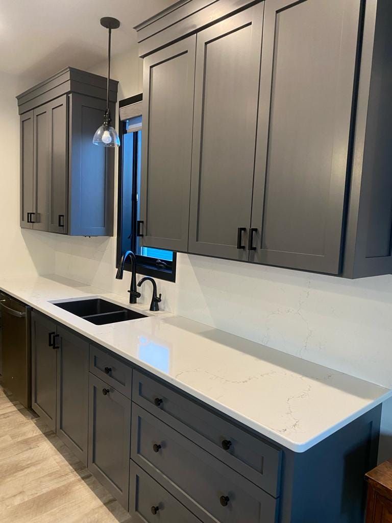 A kitchen with gray cabinets , white counter tops , and a sink.