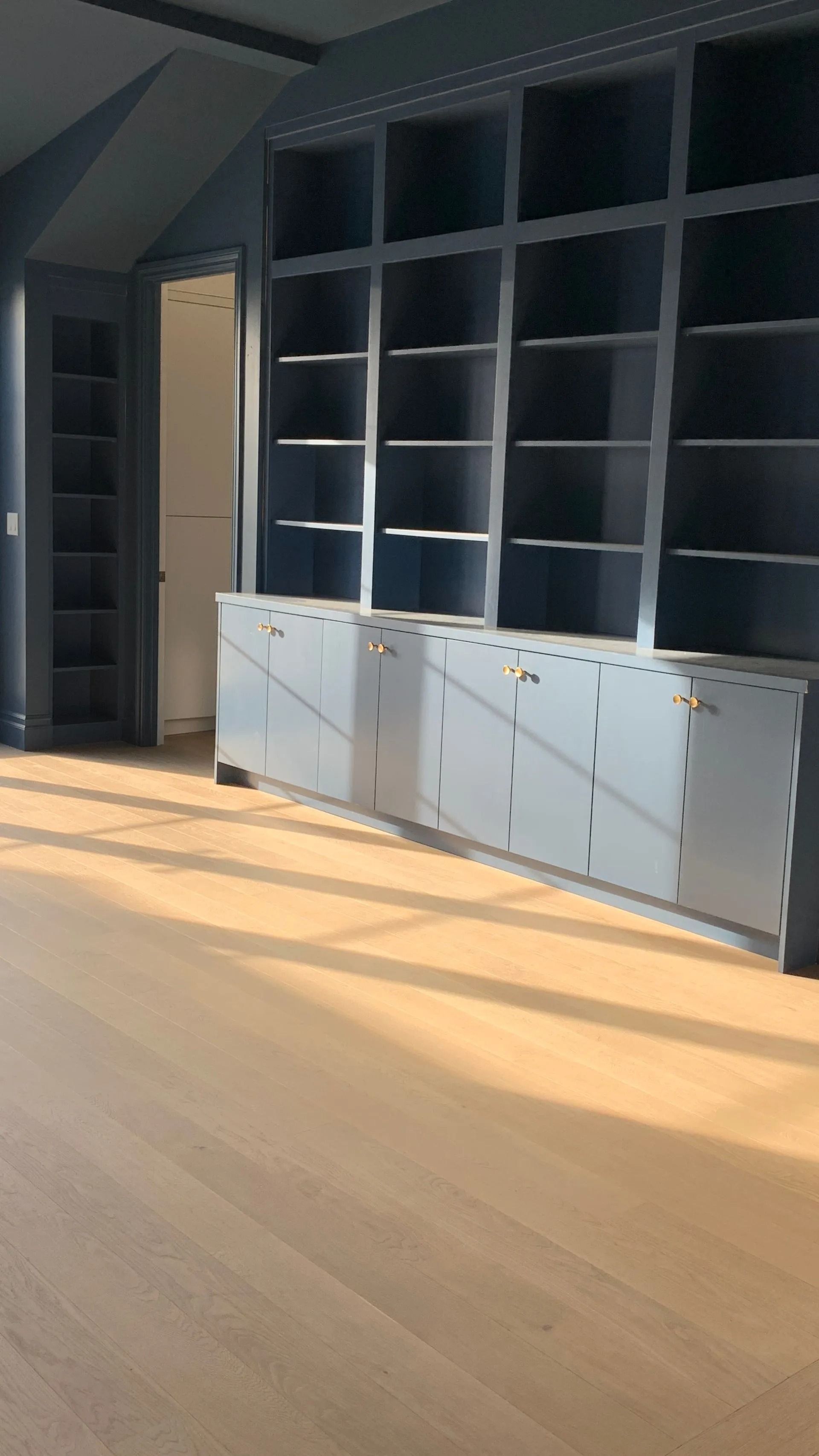 Dark blue built-in bookshelves and cabinets with light wood flooring. Sunlight streams in, illuminating the space.