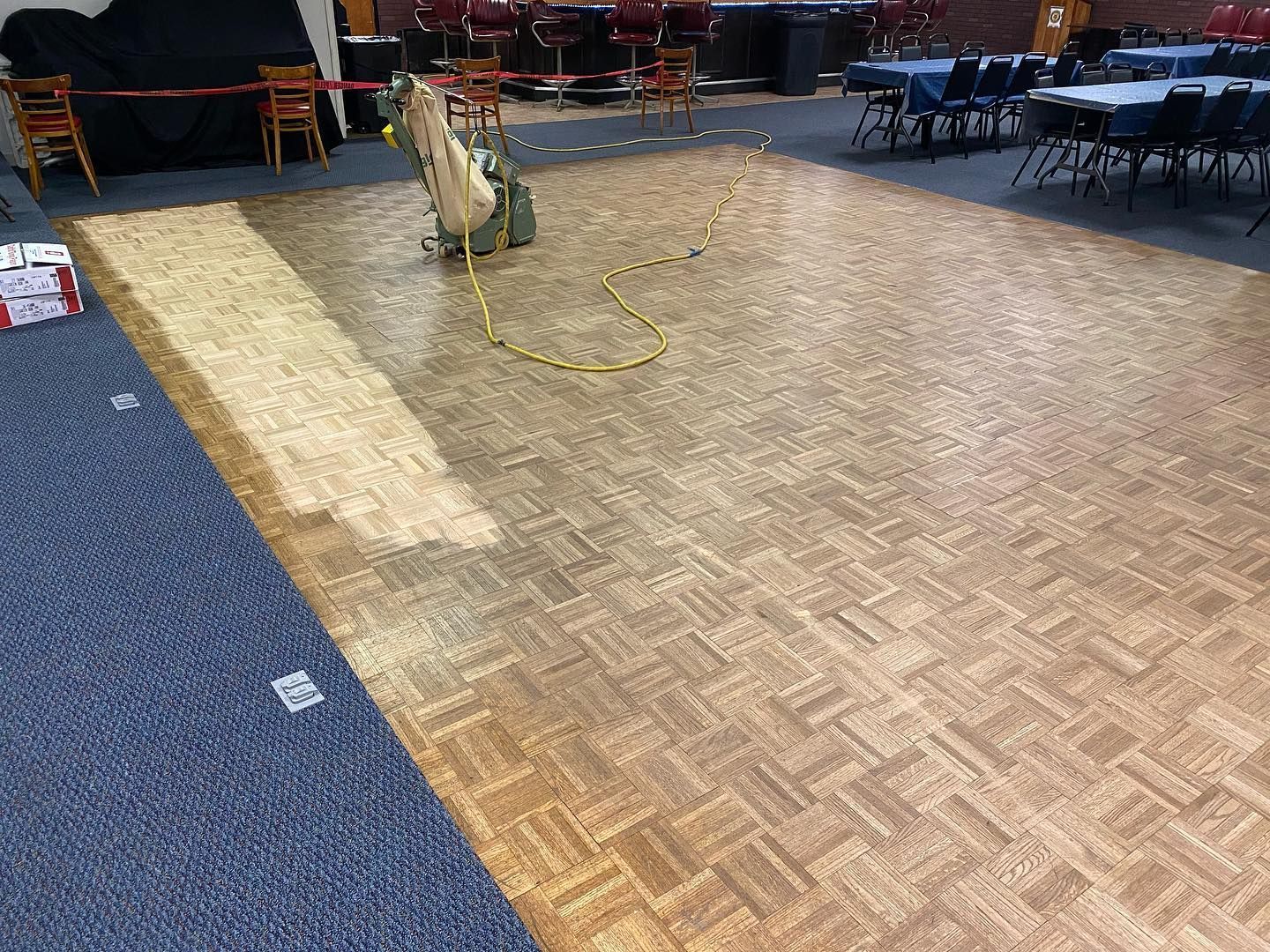 A wooden floor is being sanded in a room with tables and chairs.