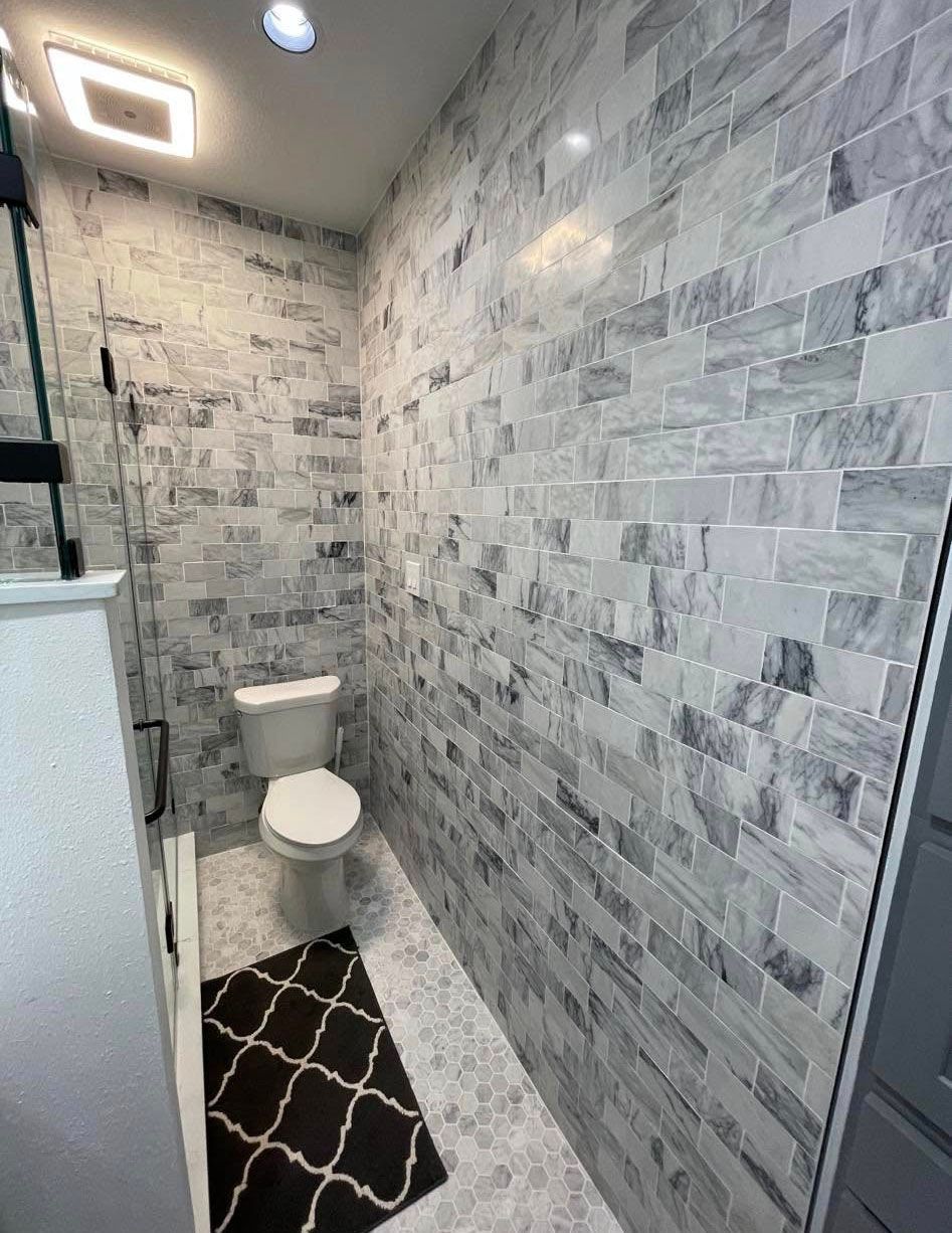 Small bathroom with marble brick-style walls, a white toilet, black rug, and a light fixture.
