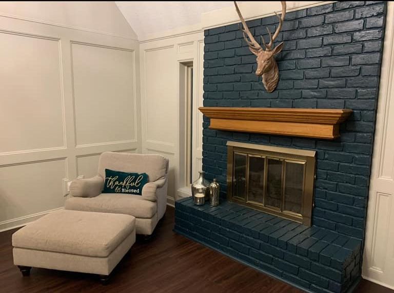 Cozy living room with a navy brick fireplace, wooden mantel, and a beige armchair with ottoman. A deer head is mounted above the fireplace.