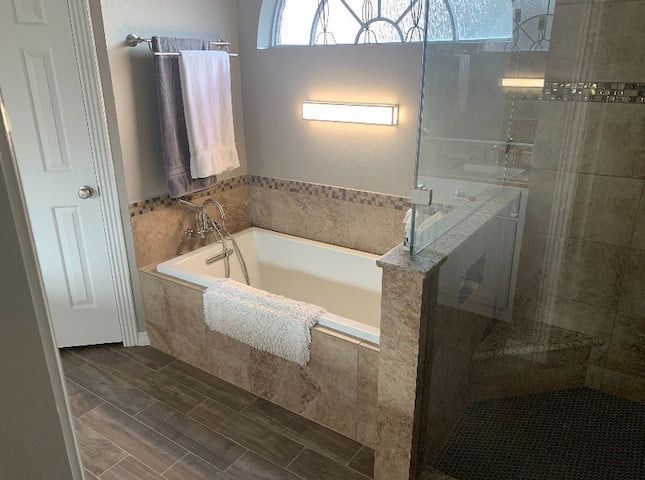 Bathroom with a built-in tub, glass shower, and wood-look tile. Beige and brown tones with towels and a light fixture.