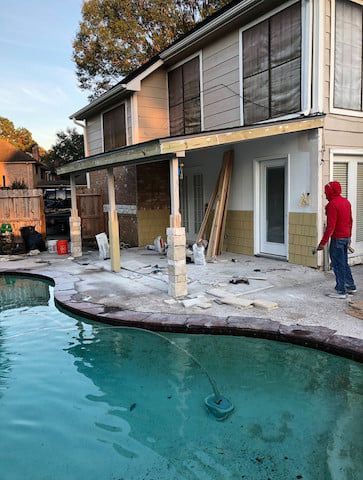 House undergoing renovation next to a pool. A person in red jacket works. Construction materials visible.