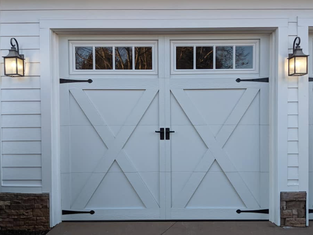 White double garage doors with black hardware and wall lanterns on a white house exterior.
