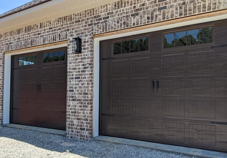 Two dark brown garage doors on a brick house exterior with a gravel driveway and blue sky reflections