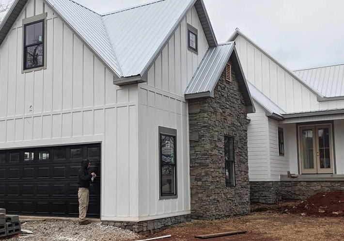 Modern white-and-stone house exterior with black garage door and metal roof under construction