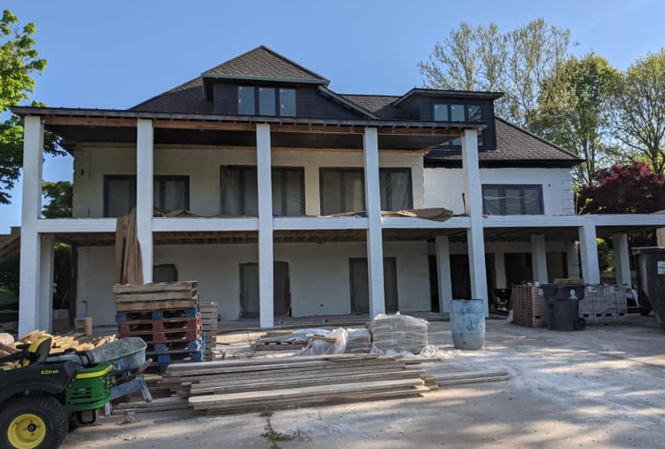 Two-story house under construction with white columns, stacked materials, and a tractor in the foreground.