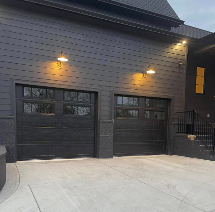 Modern gray house exterior with two black garage doors lit by wall sconces at dusk
