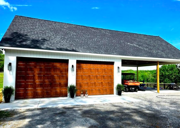 White garage with two wooden doors, gray roof, and covered carport beside a driveway under a blue sky