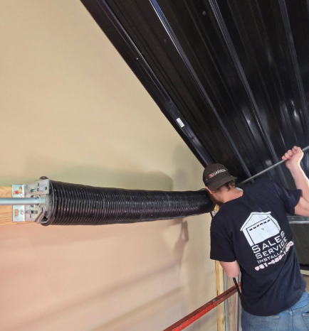 Technician adjusting a black roll-up garage door spring assembly inside a garage.