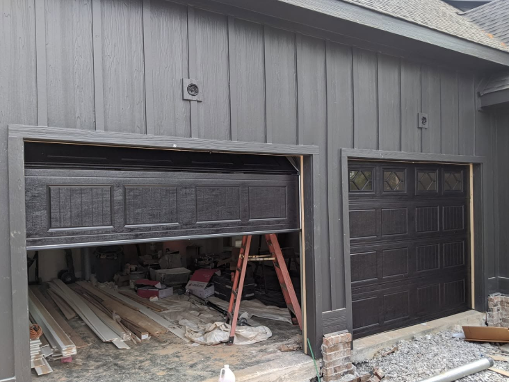 Garage exterior with one damaged, partially open door and debris inside beside a closed black garage door