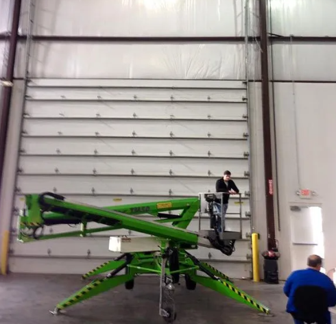 Green cherry picker lift inside a warehouse with two workers nearby and a large roll-up door