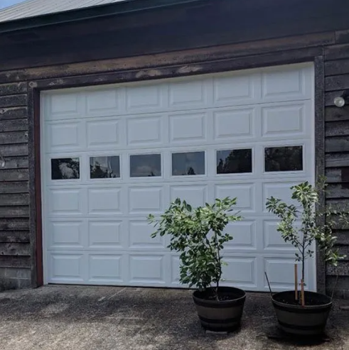White garage door on a dark wood building with two potted small trees in front