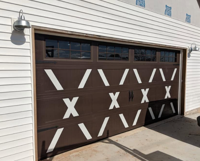 Dark brown garage door with white X-shaped tape or paint accents on a white-sided building