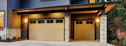 Two-car garage with tan doors, stone columns, and warm porch lights on a modern home exterior.