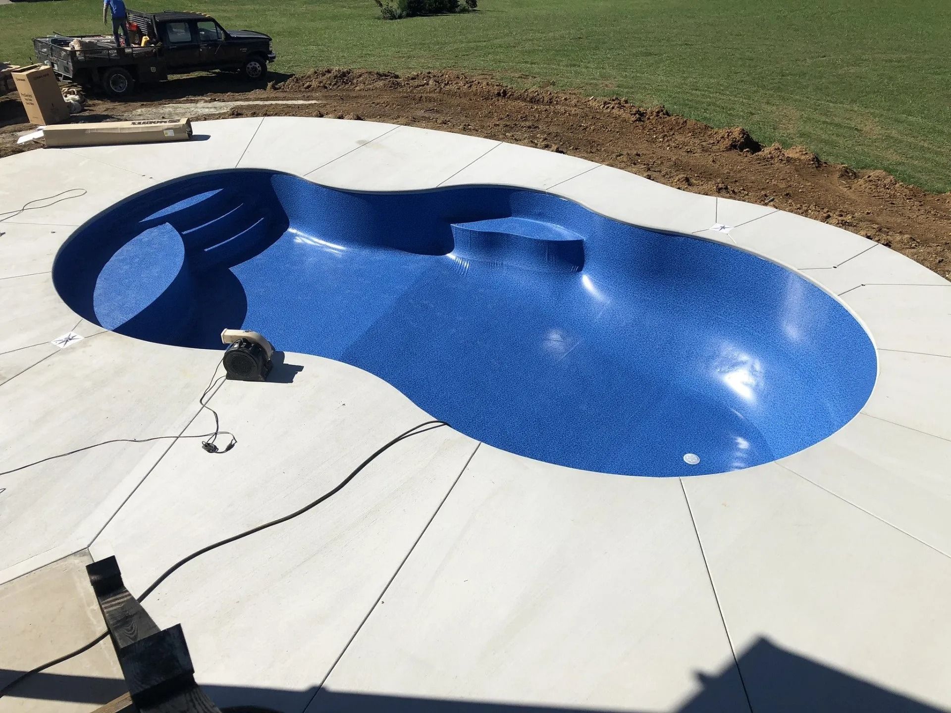 Completed blue swimming pool with concrete patio, set in a yard with a truck in the background.