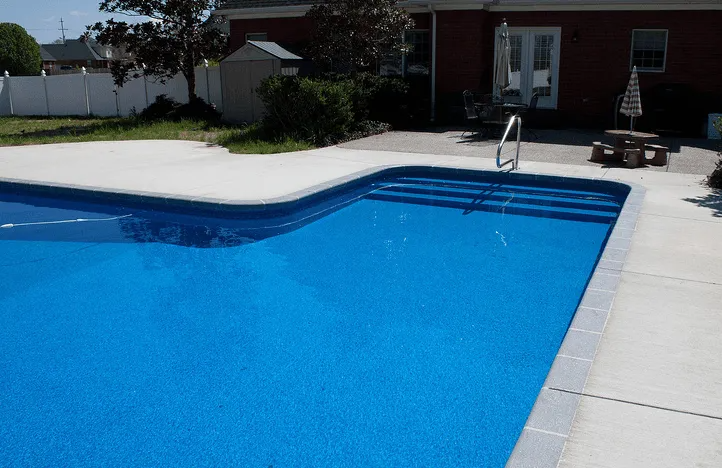 Blue swimming pool with concrete deck and steps. A house is in the background.