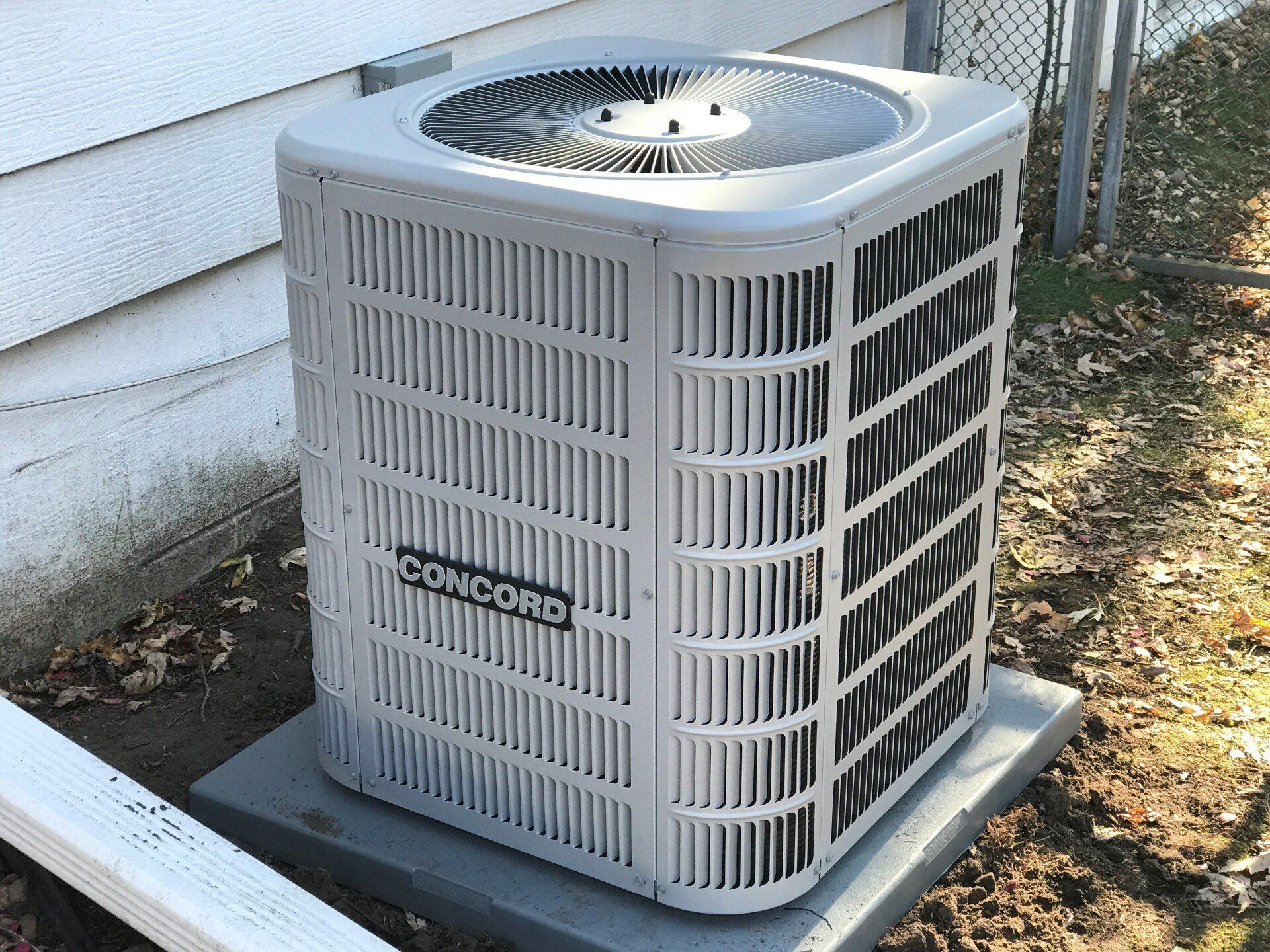 Concord air conditioning unit outdoors on a concrete pad next to a house. The unit is light gray and has a fan on top.
