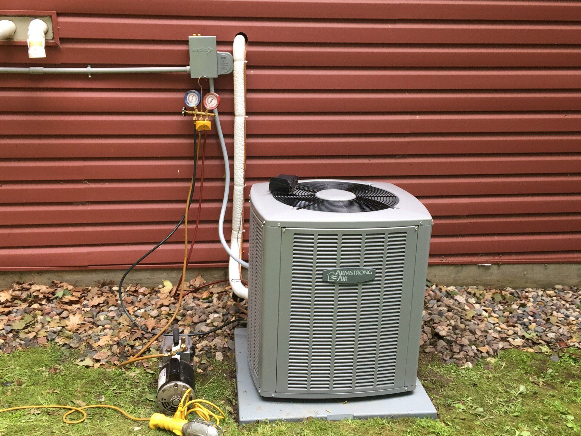 An air conditioning unit outside a building, with gauges attached, suggesting maintenance.  A vacuum pump is nearby on the grass.