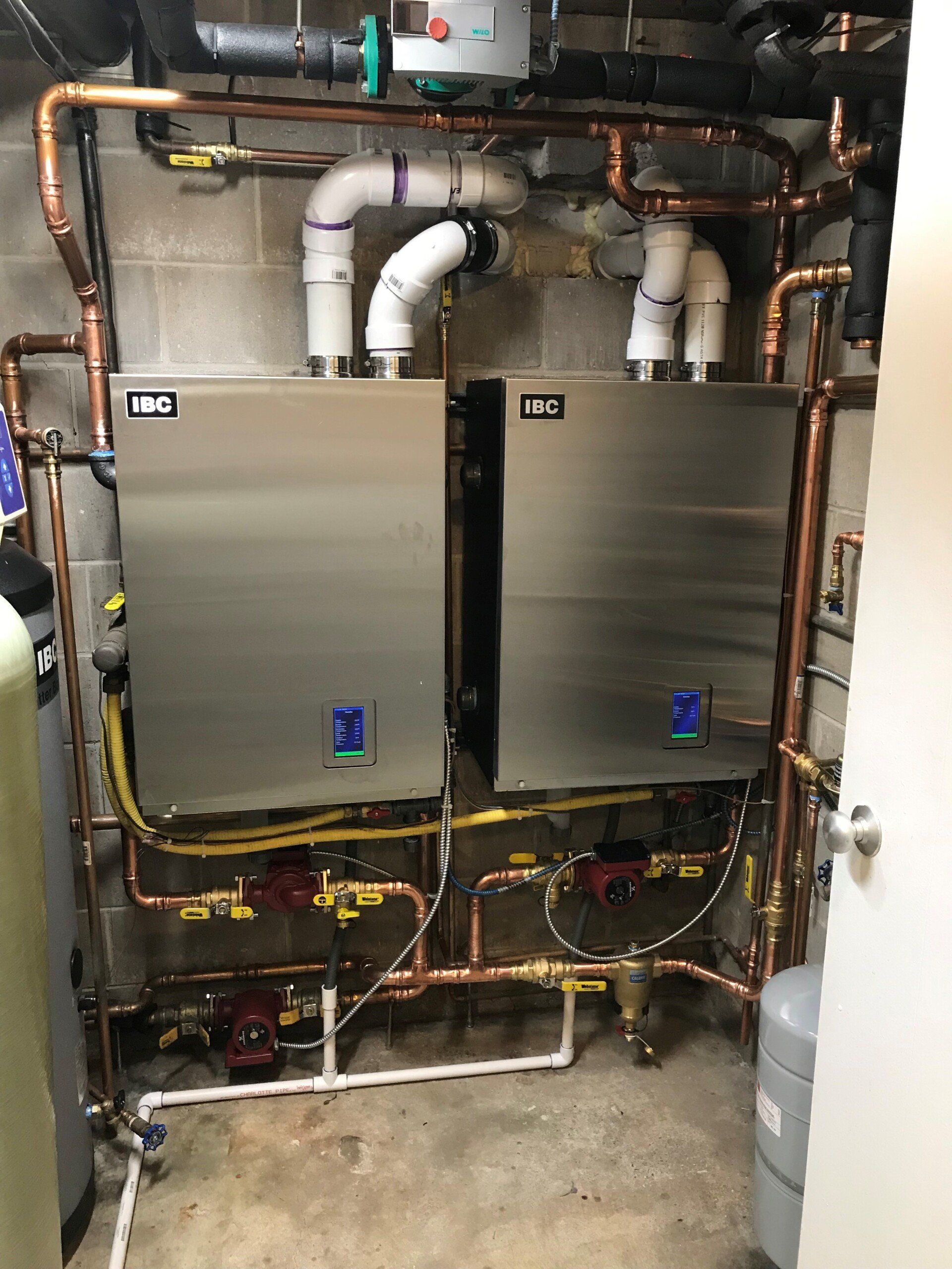 Two metallic water heaters with copper pipes in a utility room. White PVC pipes vent upwards, surrounded by brick walls.